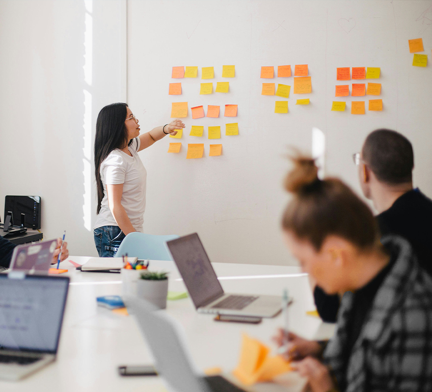 Eine junge Frau steht vor einer Wand, an der bunte Post Its hängen. Dabei zeigt und erklärt sie den Menschen, die an dem Tisch vor ihr sitzen, etwas.