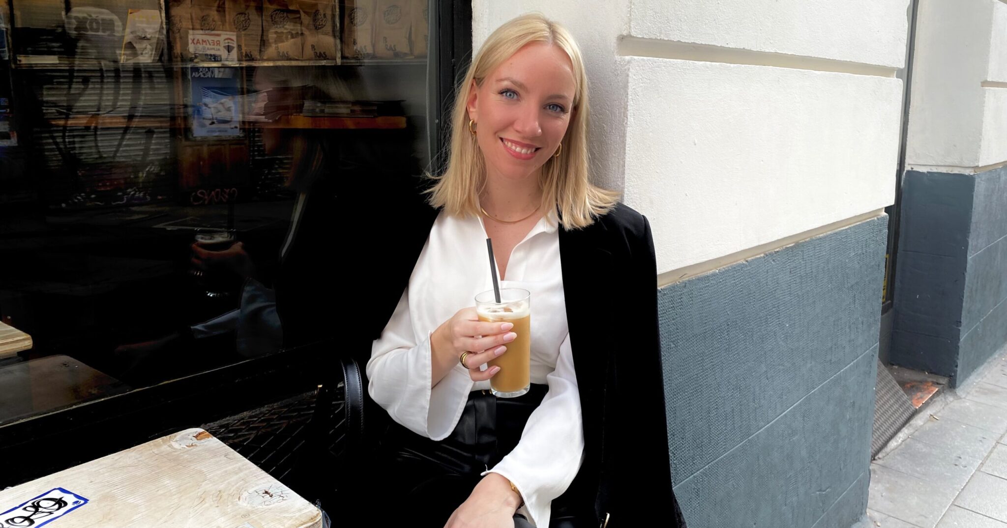 Rahel sitzt mit einem Kaffee in der Hand draußen an einem Tisch eines Cafes.